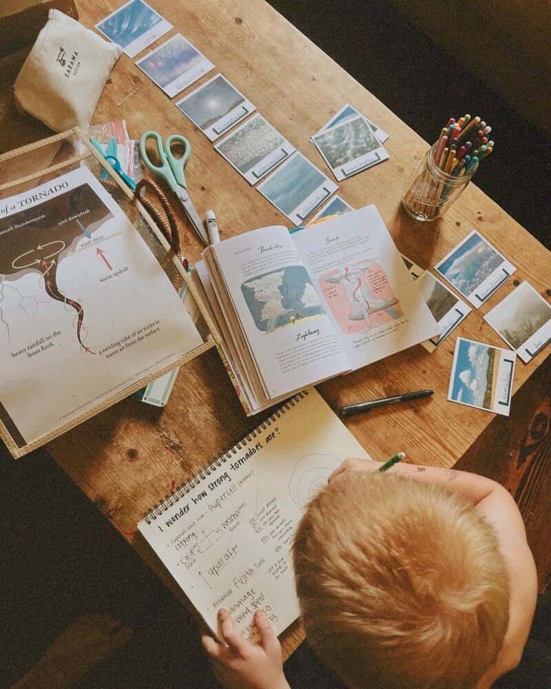 A child at a wooden table writing tornado notes beside cloud cards and a weather book